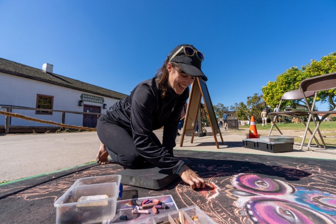 A smiling woman in black athletic wear kneels on the pavement outdoors, creating a colorful chalk art drawing.
